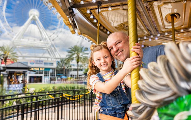 Father and daughter on a carousel at ICON Park, Orlando with The Wheel in the background.