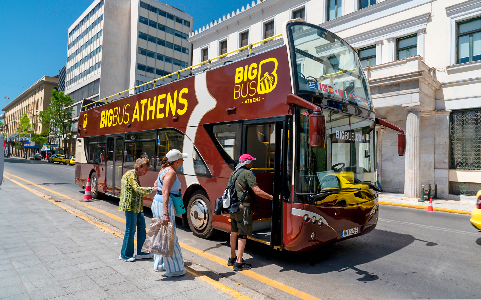 Athens hop-on hop-off bus with tourists boarding near city buildings.