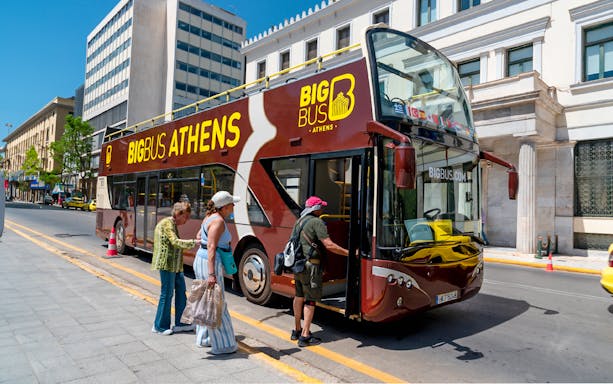 Athens hop-on hop-off bus with tourists boarding near city buildings.