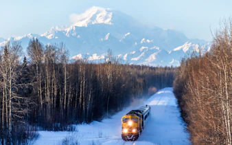 Aurora Winter Train traveling through snowy Alaskan forest with mountains in the background.