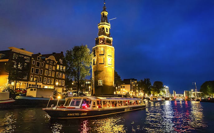 Canal cruise boat passing by Montelbaanstoren tower in Amsterdam during evening.