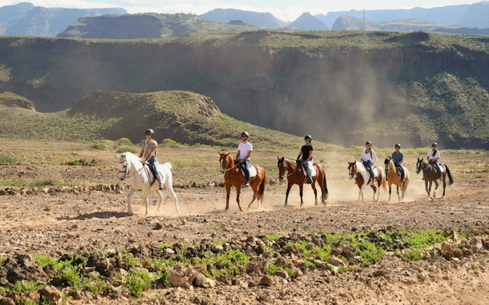Horseback riders on a volcanic trail in Gran Canaria with mountainous backdrop.