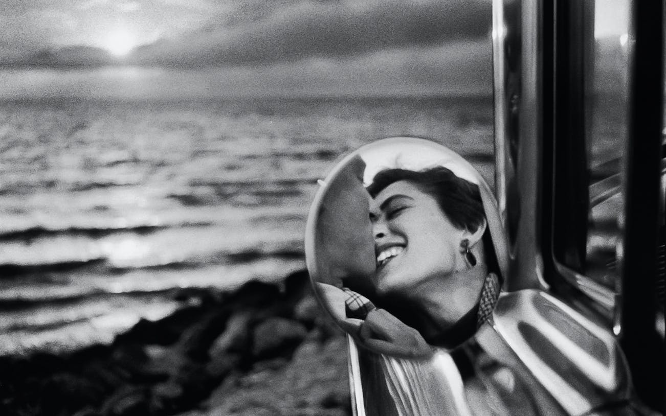 Reflection of a smiling person in a car mirror with a sunset over the ocean in the background.
