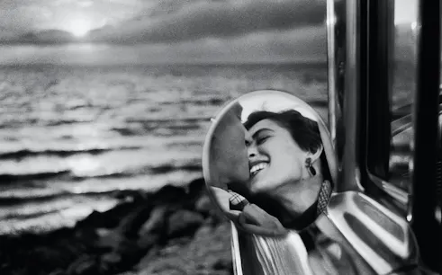 Reflection of a smiling person in a car mirror with a sunset over the ocean in the background.