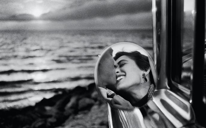 Reflection of a smiling person in a car mirror with a sunset over the ocean in the background.