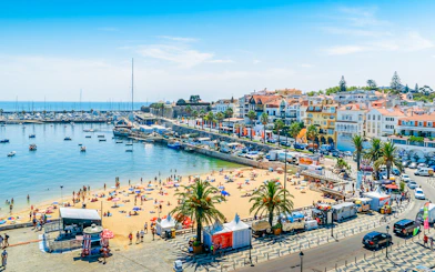 Crowded Praia do Ribeiro beach with boats in the bay on a sunny day in Cascais, Portugal.