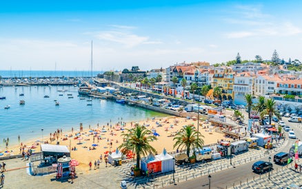 Crowded Praia do Ribeiro beach with boats in the bay on a sunny day in Cascais, Portugal.