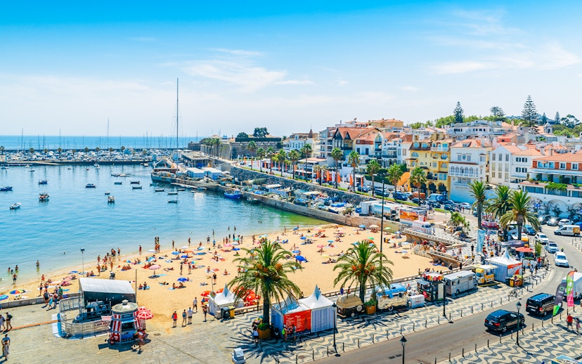Crowded Praia do Ribeiro beach with boats in the bay on a sunny day in Cascais, Portugal.