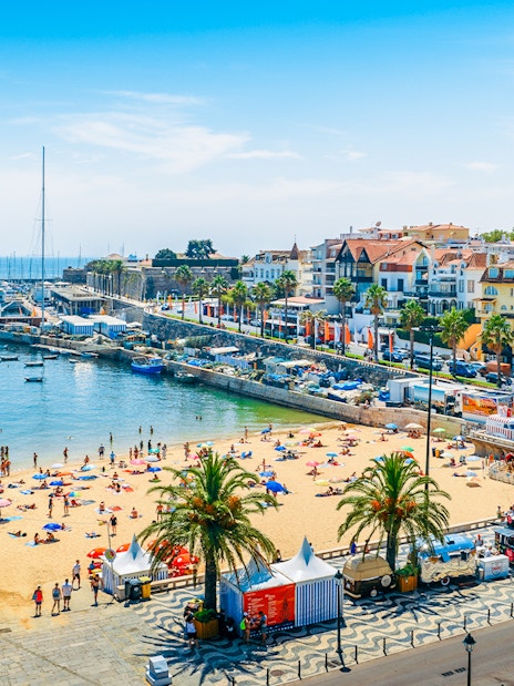 Crowded Praia do Ribeiro beach with boats in the bay on a sunny day in Cascais, Portugal.