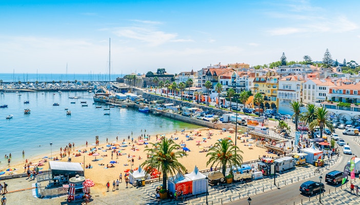 Crowded Praia do Ribeiro beach with boats in bay, sunny day, Cascais, Portugal, near Lisbon.