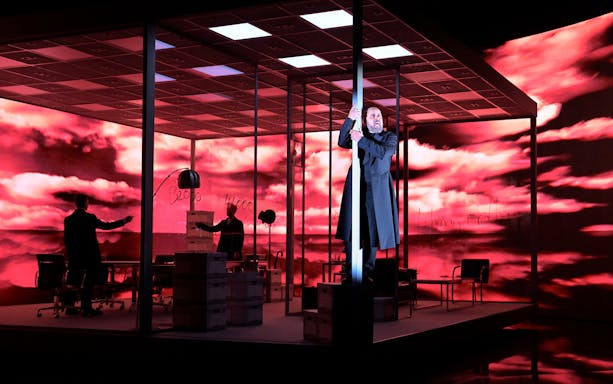 Stage scene from The Lehman Trilogy with actors in an office setting against a dramatic red sky backdrop.