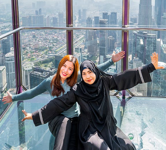 Tourists posing in the Kuala Lumpur Tower Glass Box with city skyline view.