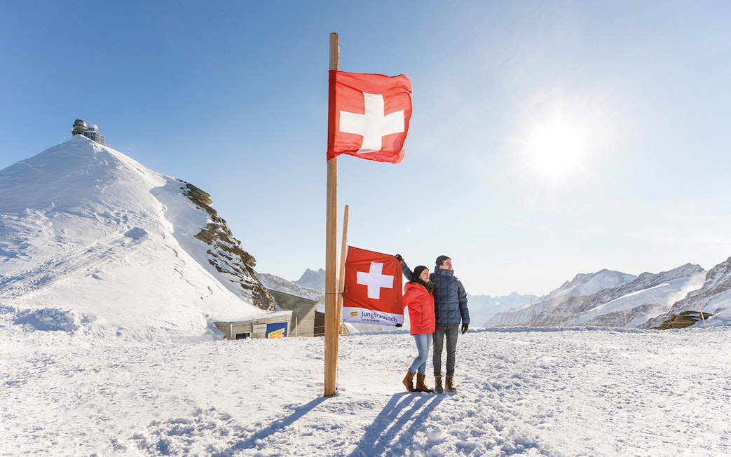 Couple standing by Swiss flags at snowy Jungfraujoch, with observatory and mountains in background.