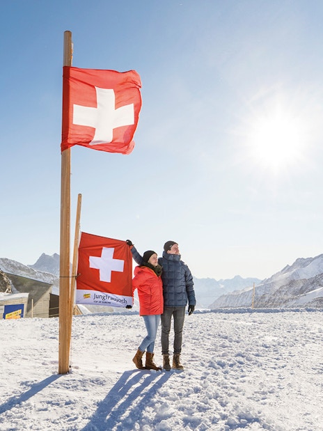 Couple standing by Swiss flags at snowy Jungfraujoch, with observatory and mountains in background.
