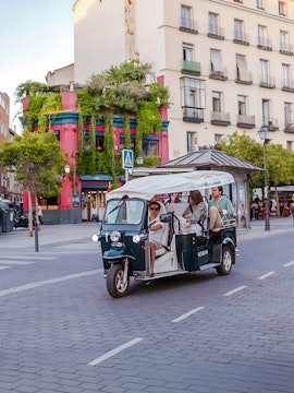 Electric tuk-tuk tour passing colorful building in city street.