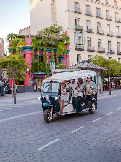 Electric tuk-tuk tour passing colorful building in city street.
