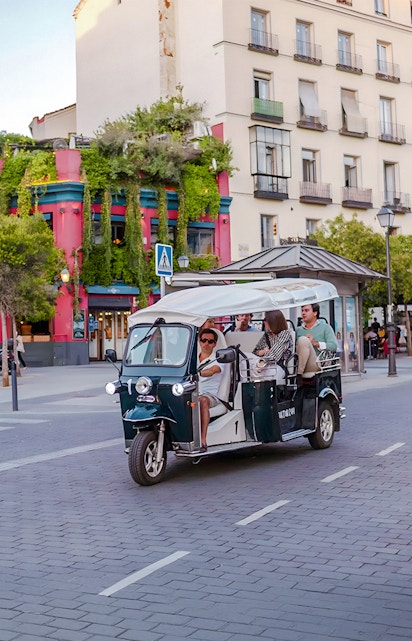 Electric tuk-tuk tour passing colorful building in city street.