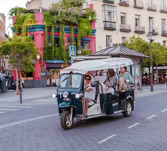 Electric tuk-tuk tour passing colorful building in city street.