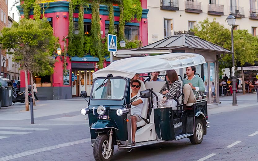 Electric tuk-tuk tour passing colorful building in city street.