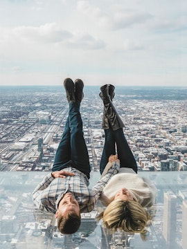 Couple lying on glass ledge at SkyDeck, Willis Tower, overlooking Chicago skyline.