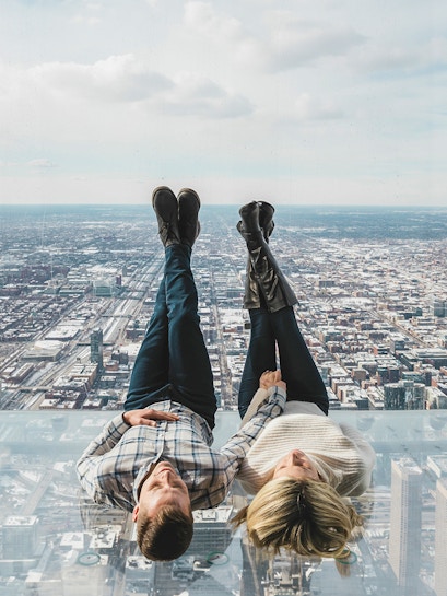 Couple lying on glass ledge at SkyDeck, Willis Tower, overlooking Chicago skyline.