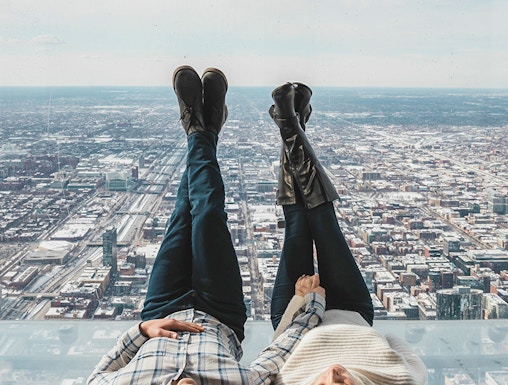 Couple lying on glass ledge at SkyDeck, Willis Tower, overlooking Chicago skyline.
