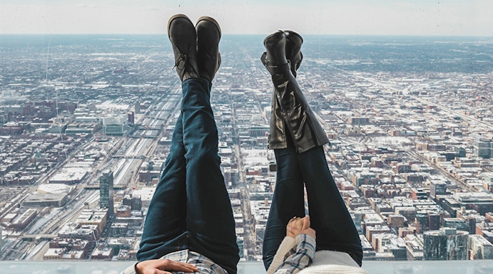 Couple lying on glass ledge at SkyDeck, Willis Tower, overlooking Chicago skyline.