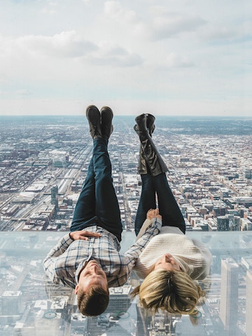 Couple lying on glass ledge at SkyDeck, Willis Tower, overlooking Chicago skyline.