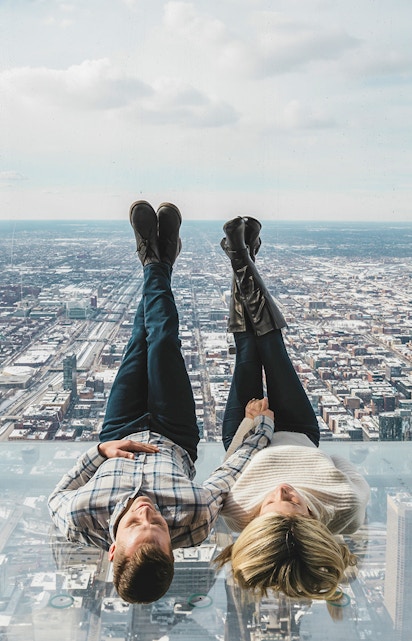 Couple lying on glass ledge at SkyDeck, Willis Tower, overlooking Chicago skyline.