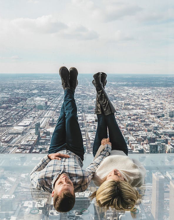 Couple lying on glass ledge at SkyDeck, Willis Tower, overlooking Chicago skyline.
