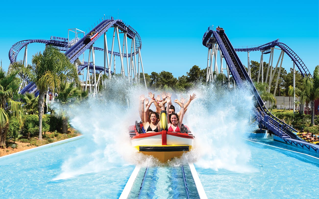 Visitors enjoying a water ride at Aquashow Water Park with roller coasters in the background.