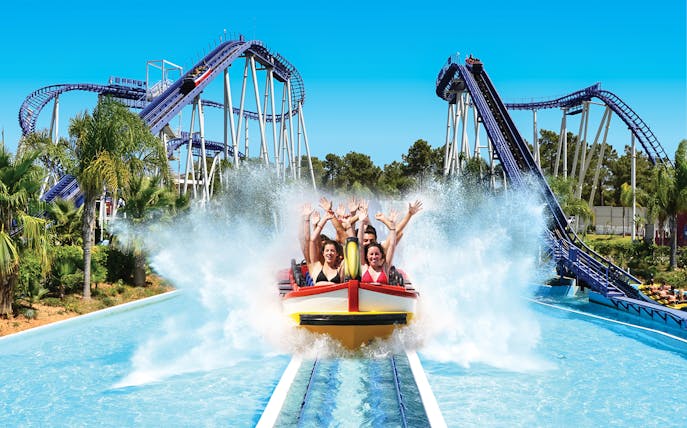 Visitors enjoying a water ride at Aquashow Water Park with roller coasters in the background.