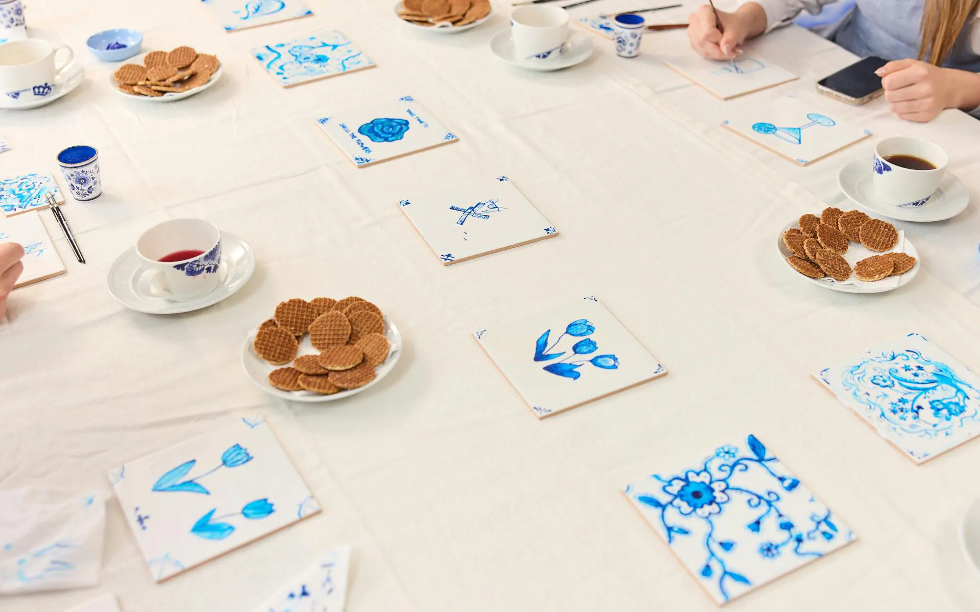 Guests painting traditional Dutch blue tiles at a workshop, with tea and stroopwafels on the table.