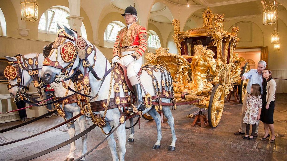 Royal carriage with ornate gold details and tourists observing at Buckingham Palace.