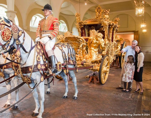 Royal carriage with ornate gold details and tourists observing at Buckingham Palace.