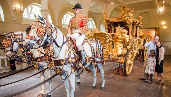 Royal carriage with ornate gold details and tourists observing at Buckingham Palace.