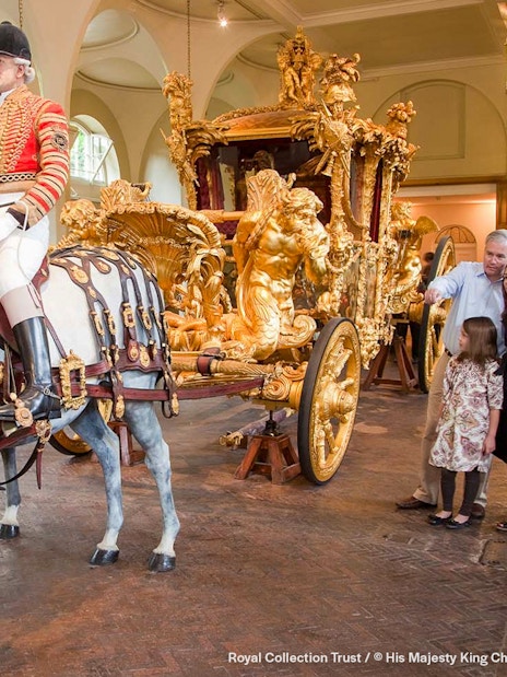 Royal carriage with ornate gold details and tourists observing at Buckingham Palace.