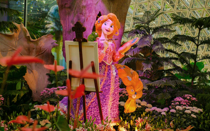 Rapunzel painting in Disney Garden of Wonder surrounded by vibrant flowers and greenery.
