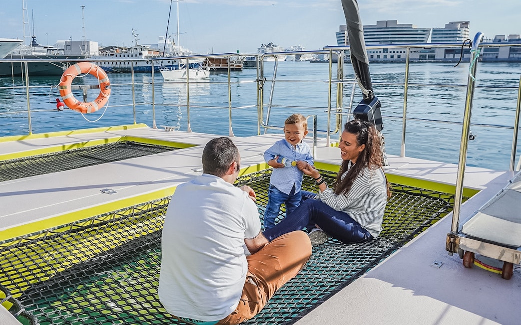 Tourists relaxing on catamaran cruise deck with child, harbor view in background.