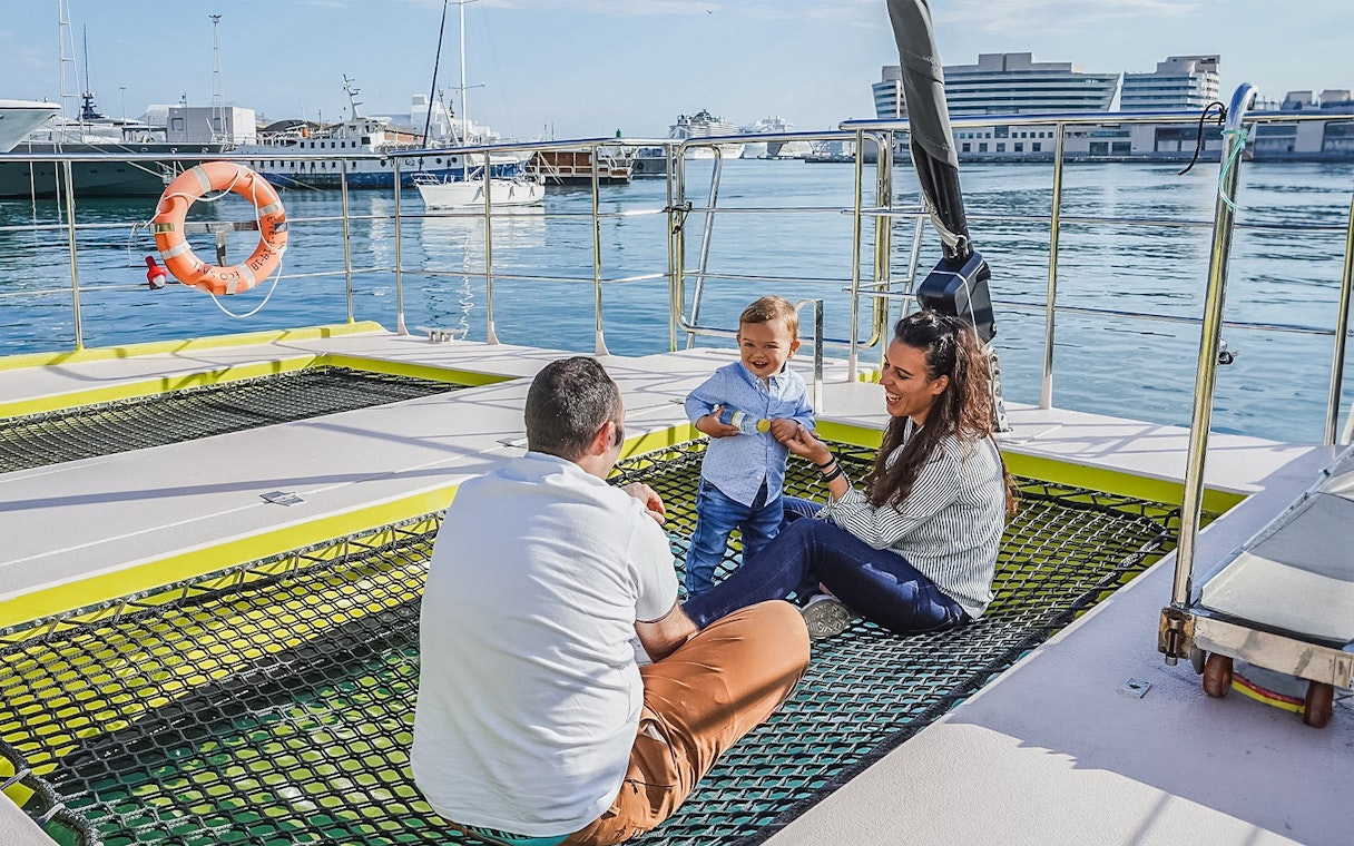 Tourists relaxing on catamaran cruise deck with child, harbor view in background.