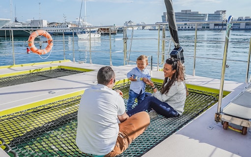 Tourists relaxing on catamaran cruise deck with child, harbor view in background.