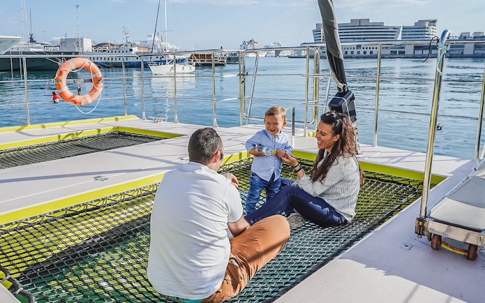 Tourists relaxing on catamaran cruise deck with child, harbor view in background.
