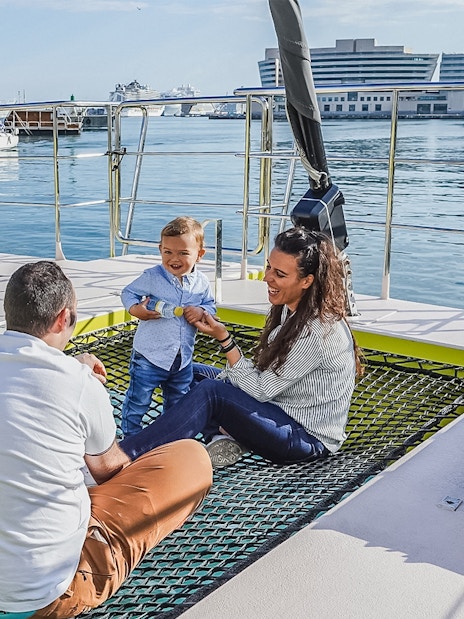 Tourists relaxing on catamaran cruise deck with child, harbor view in background.