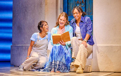 Three actors in a scene from Mamma Mia, sitting on steps with a book, in front of a blue door.