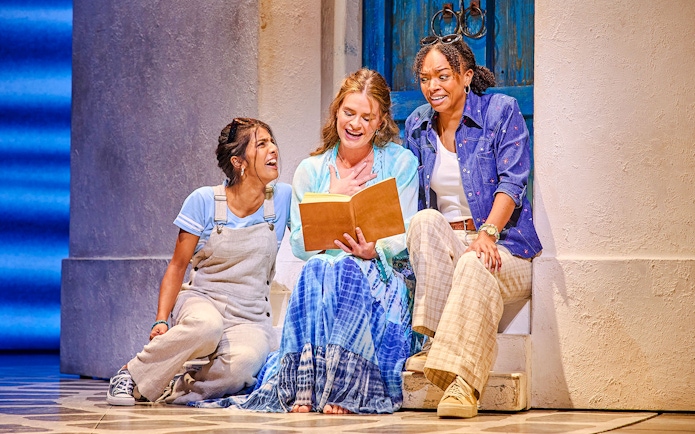 Three actors in a scene from Mamma Mia, sitting on steps with a book, in front of a blue door.