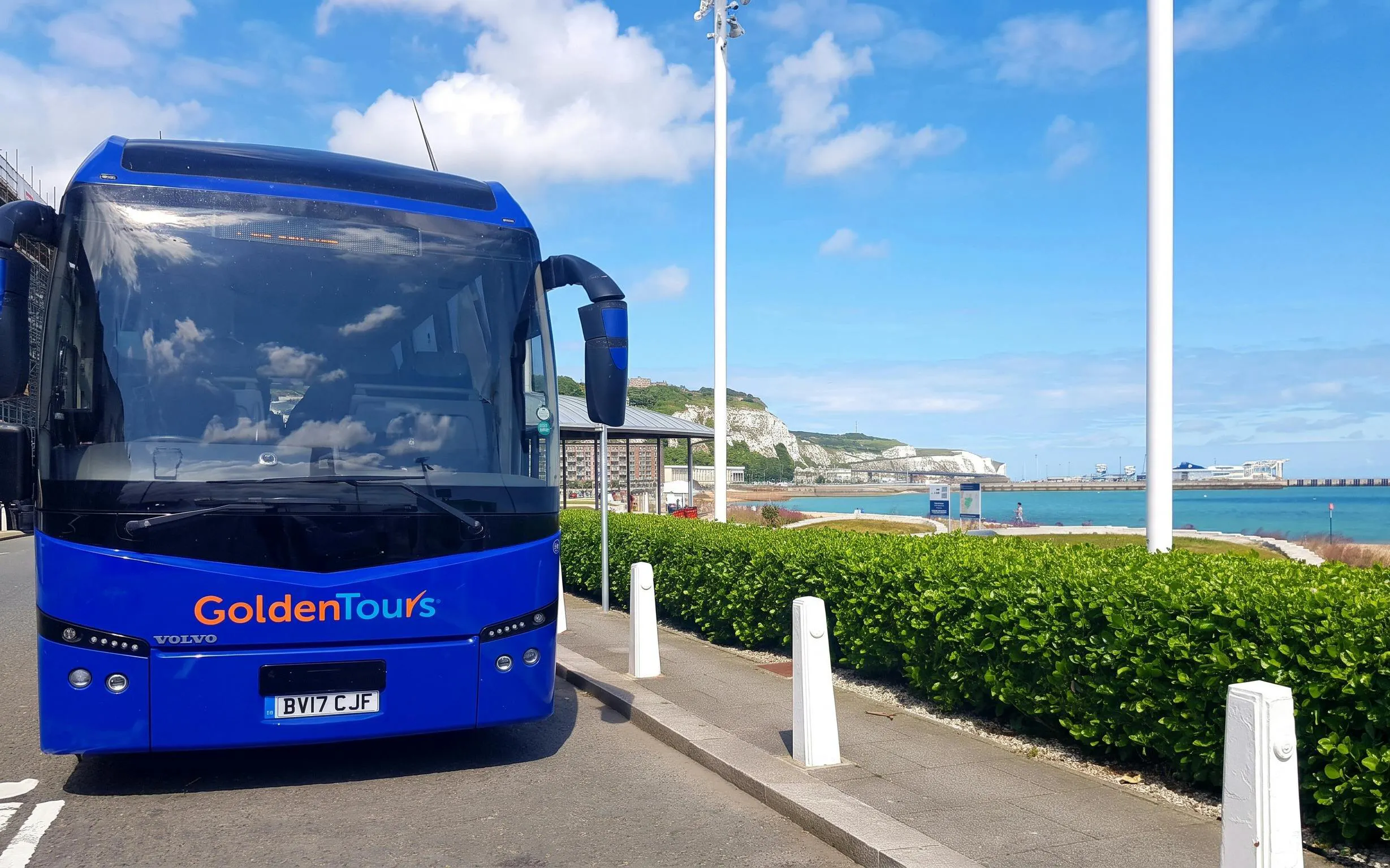 AC coach parked near coastal road with sea and cliffs in the background.
