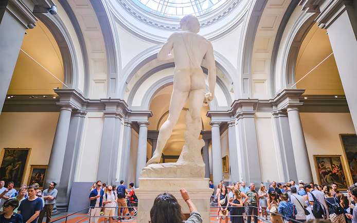 David statue's back view in Accademia Gallery, Florence, with visitors observing.