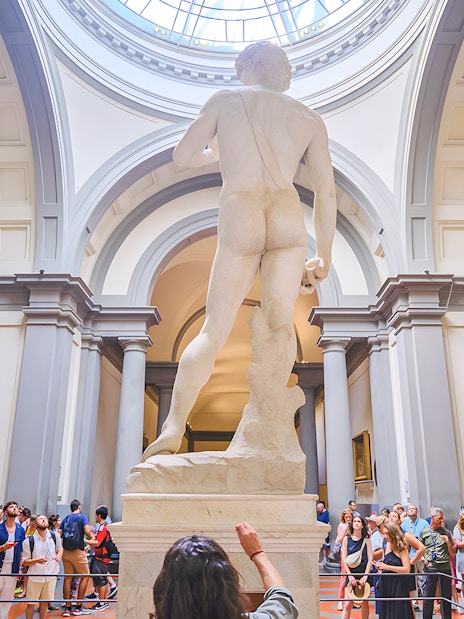 David statue's back view in Accademia Gallery, Florence, with visitors observing.