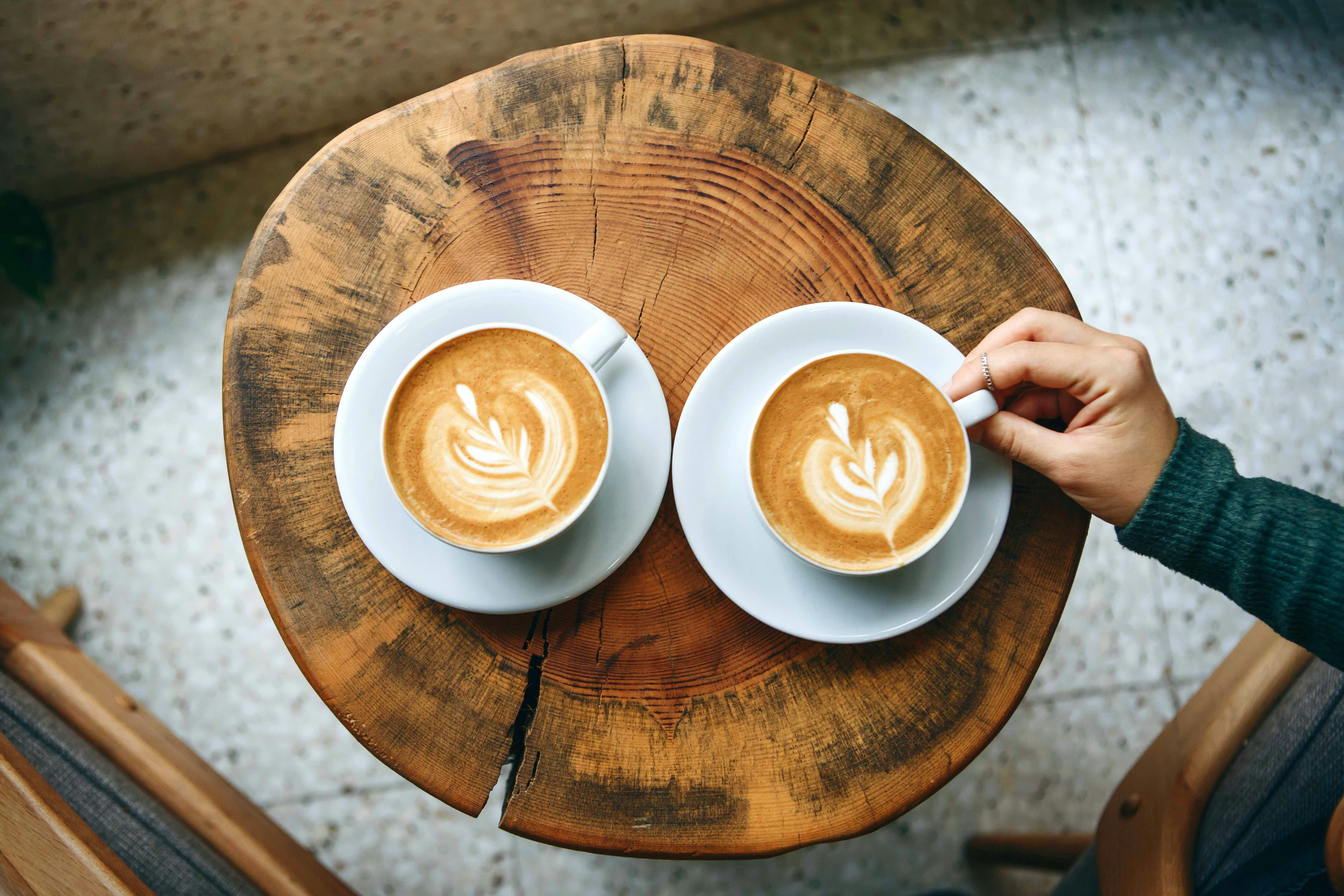 Two cups of latte with latte art on a wooden table, one being held by a hand.