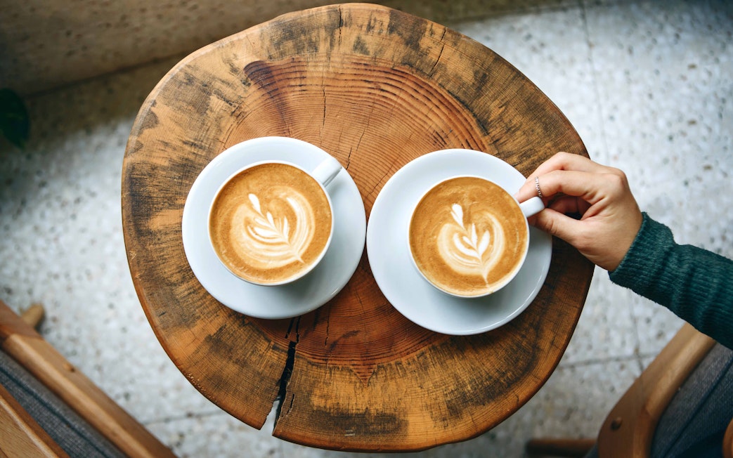 Two cups of latte with latte art on a wooden table, one being held by a hand.